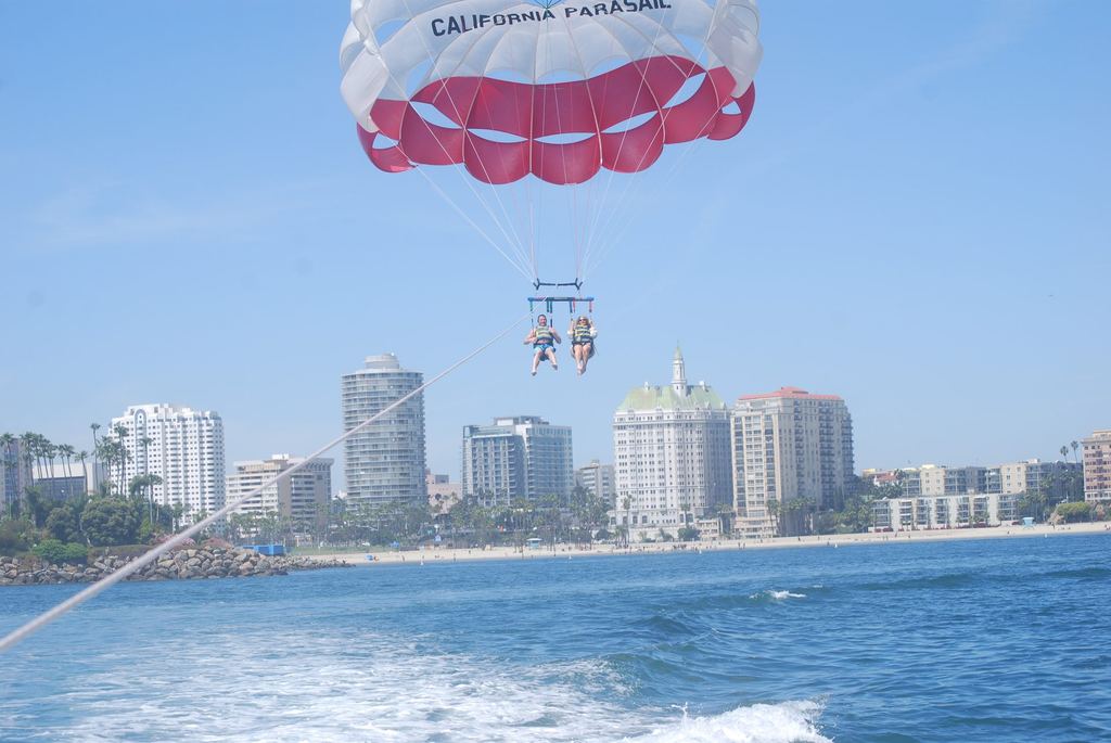 Two flyers side by side under a red and white parasail above the water off Long Beach, with the downtown skyline, palm trees and the beach behind them