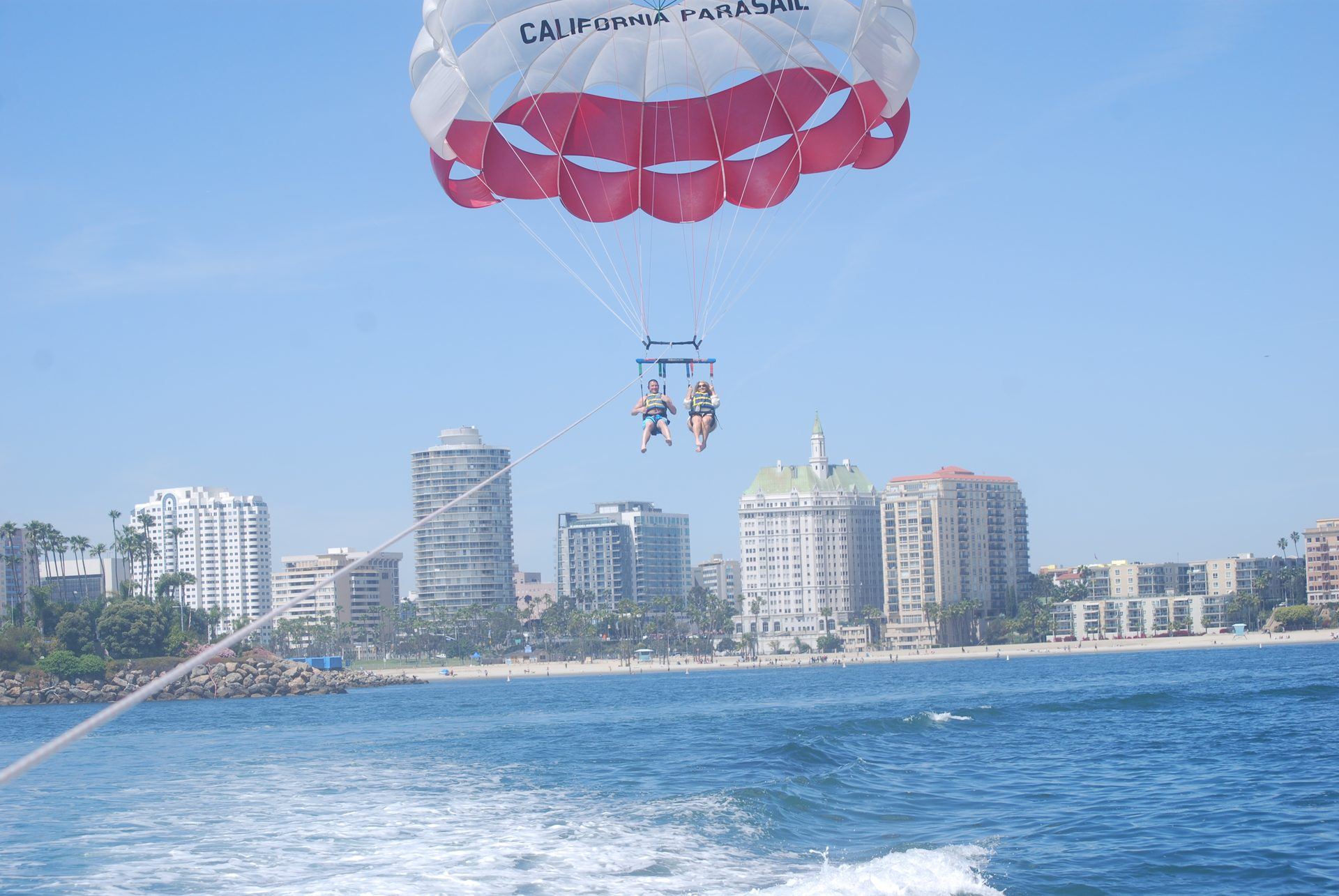 Two flyers side by side under a red and white parasail above the water off Long Beach, with the downtown skyline, palm trees and the beach behind them
